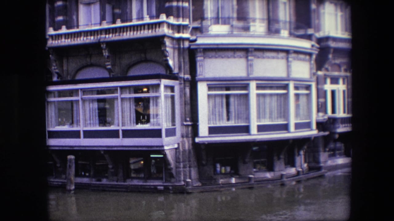 1967 Amsterdam canal-side building facade seen from water level, with ornate masonry, bay windows, curtains, balcony railings, and canal water in the foreground.