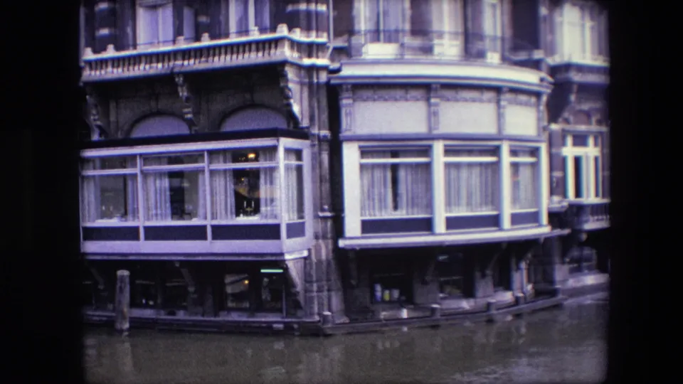 1967 Amsterdam canal-side building facade seen from water level, with ornate masonry, bay windows, curtains, balcony railings, and canal water in the foreground.
