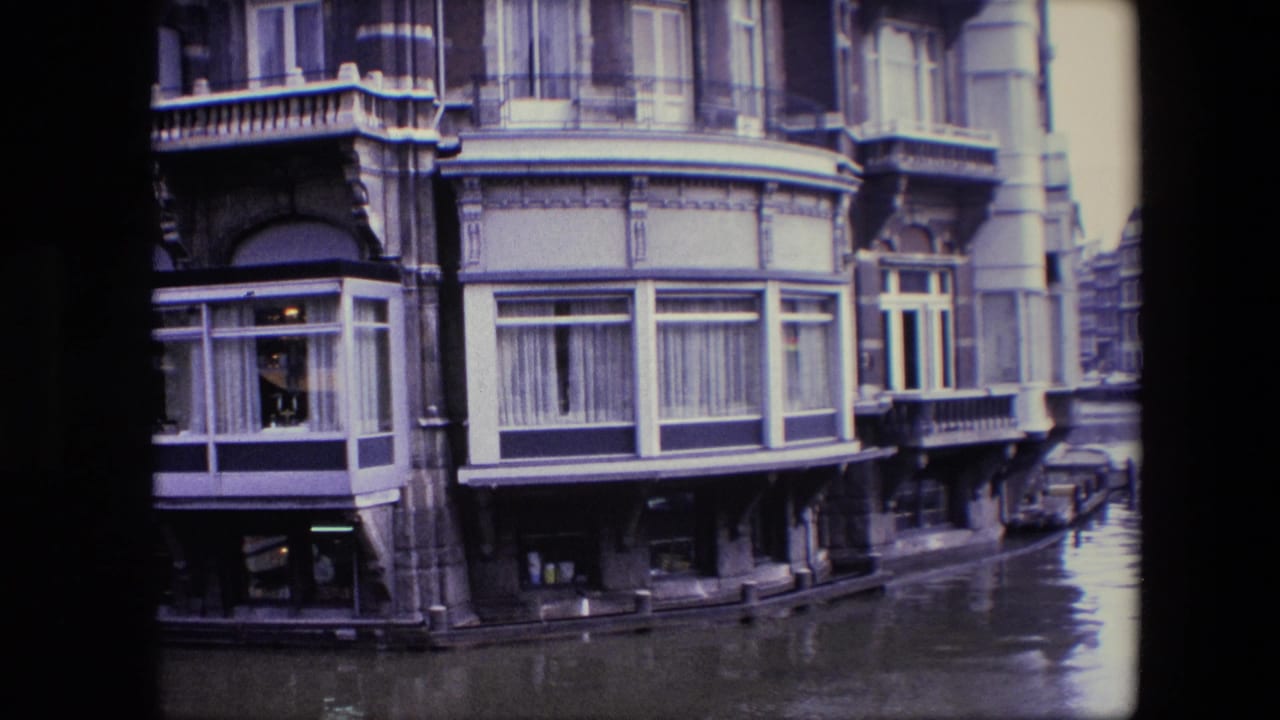 1967 Amsterdam canal-front building corner with large bay windows, balcony trim, water below, and a narrow street or canal edge visible to the right.
