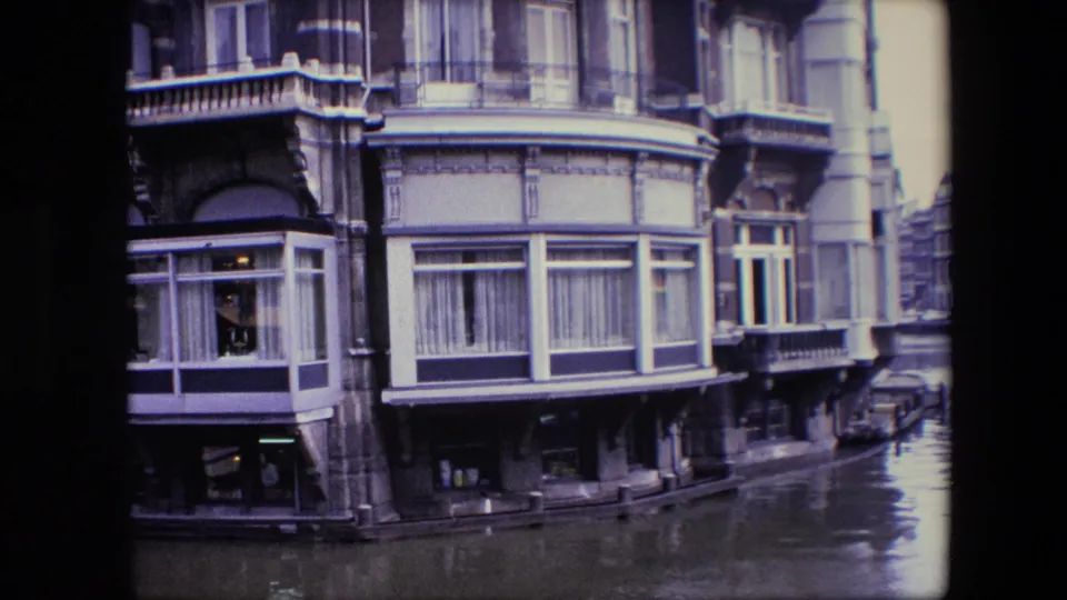 1967 Amsterdam canal-front building corner with large bay windows, balcony trim, water below, and a narrow street or canal edge visible to the right.