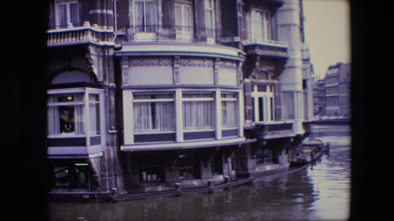 1967 Amsterdam canal-side facade with bay windows and balcony details, canal water in front, and a parked car partly visible near the street edge.