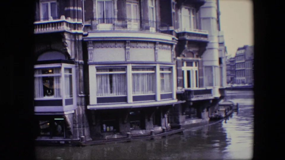 1967 Amsterdam canal-side facade with bay windows and balcony details, canal water in front, and a parked car partly visible near the street edge.