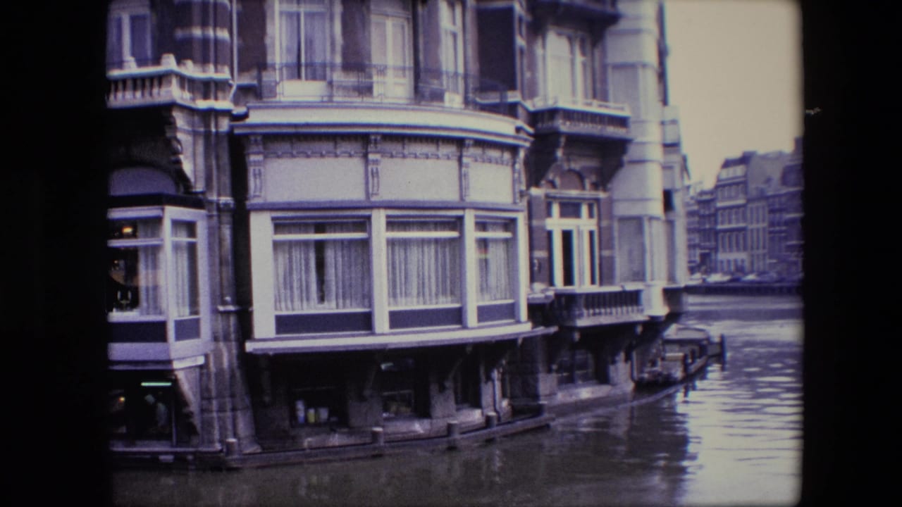 1967 home-movie frame of an ornate Amsterdam canal-side building with bay windows, curtains, balconies, and water reflecting the facade.