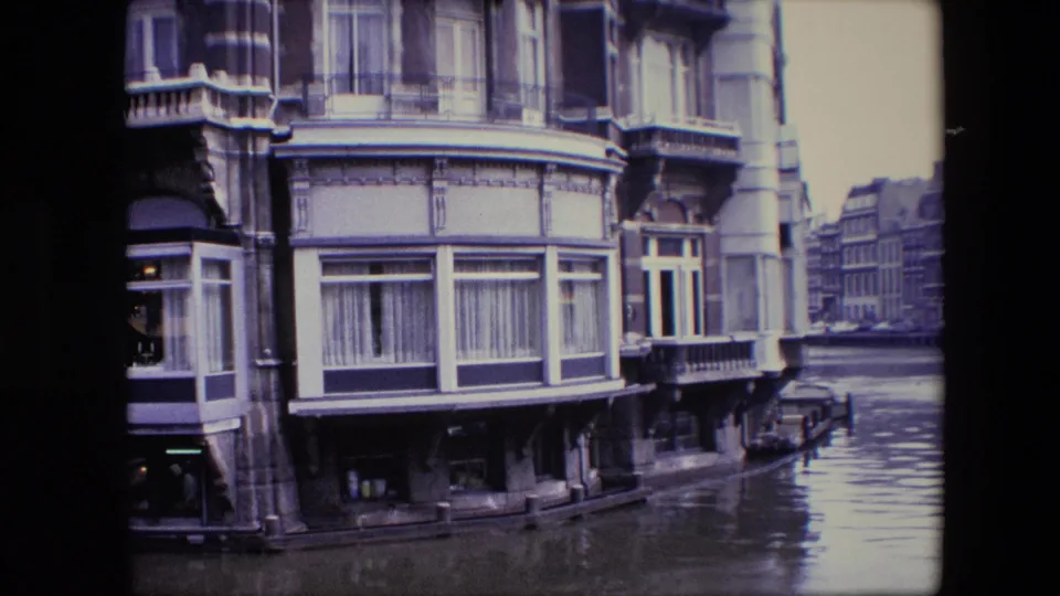 1967 home-movie frame of an ornate Amsterdam canal-side building with bay windows, curtains, balconies, and water reflecting the facade.