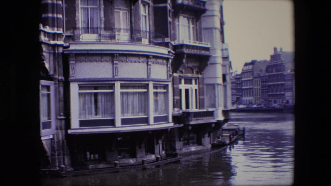 Amsterdam canal view showing a multi-story historic facade at the waterline with balcony details and more buildings receding along the canal.