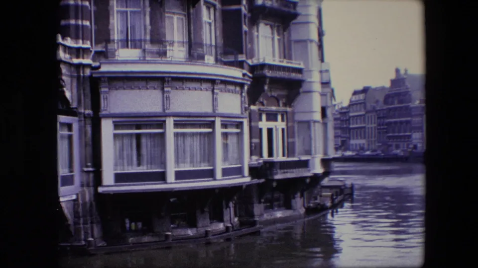 Amsterdam canal view showing a multi-story historic facade at the waterline with balcony details and more buildings receding along the canal.
