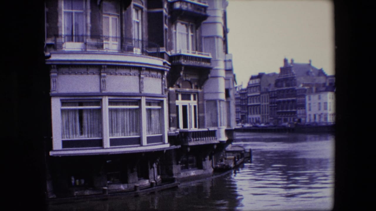 Mid-century 8mm frame of Amsterdam canal architecture with a close facade, curtained windows, decorative stonework, and rippled canal water below.