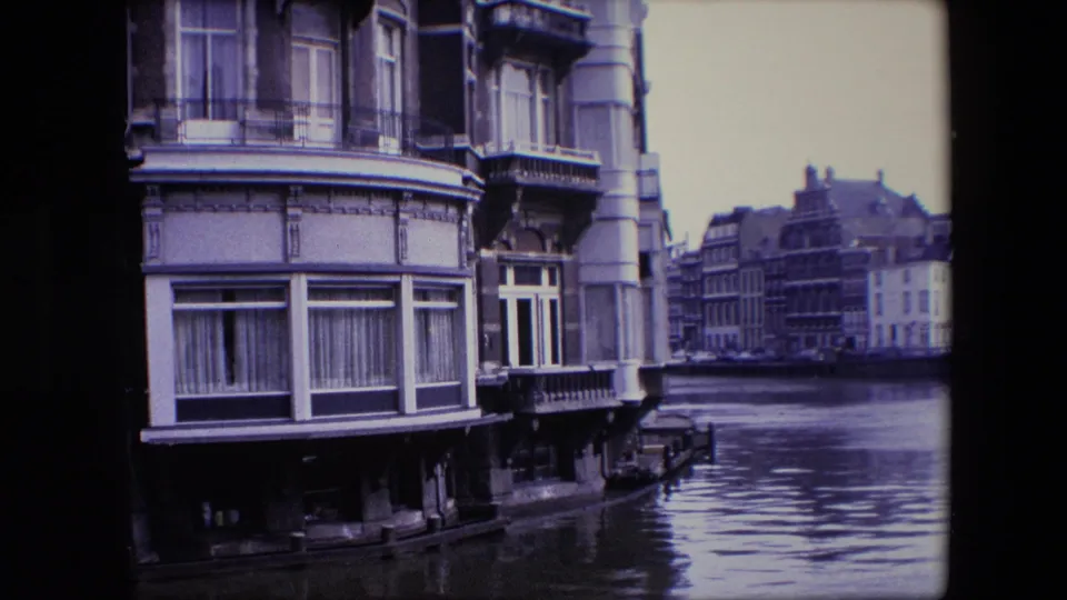 Mid-century 8mm frame of Amsterdam canal architecture with a close facade, curtained windows, decorative stonework, and rippled canal water below.