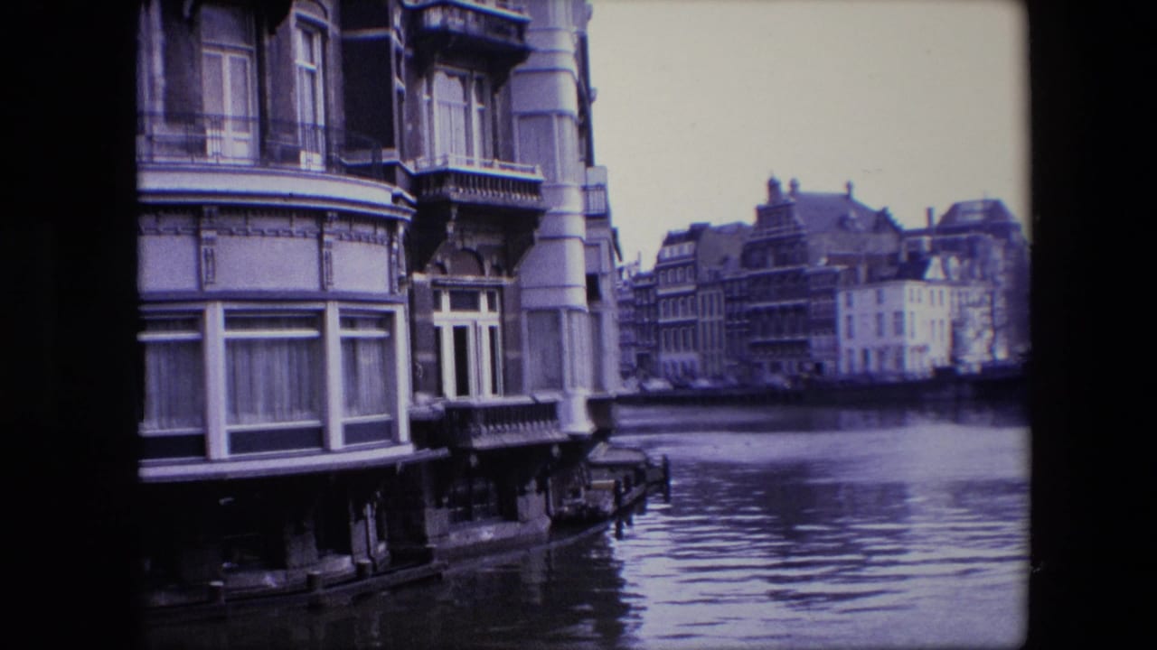 Amsterdam canal scene with the foreground building partly cropped, rippled water across the lower frame, and historic waterfront facades in the distance.