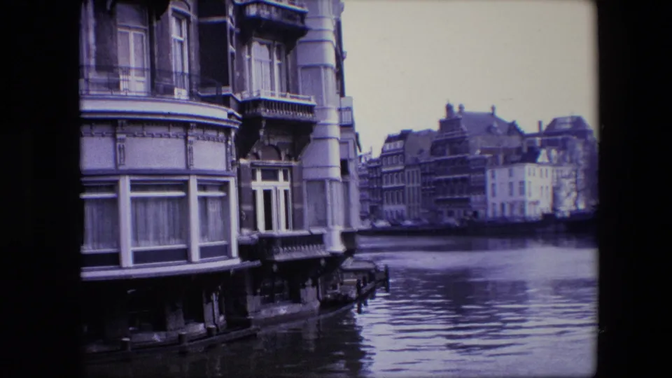 Amsterdam canal scene with the foreground building partly cropped, rippled water across the lower frame, and historic waterfront facades in the distance.