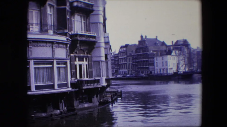 1967 Amsterdam canal view with ornate waterfront buildings, tall windows, decorative columns, and reflections across the water.