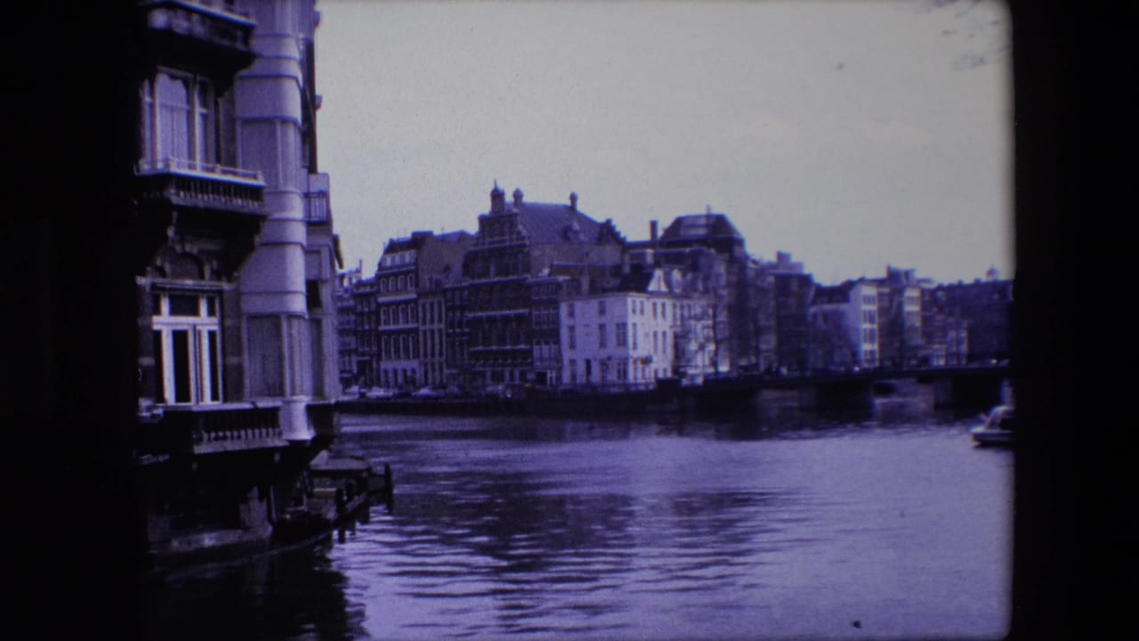 1967 Amsterdam canal scene with a close architectural edge, older waterfront buildings across the canal, and rippled water filling the foreground.