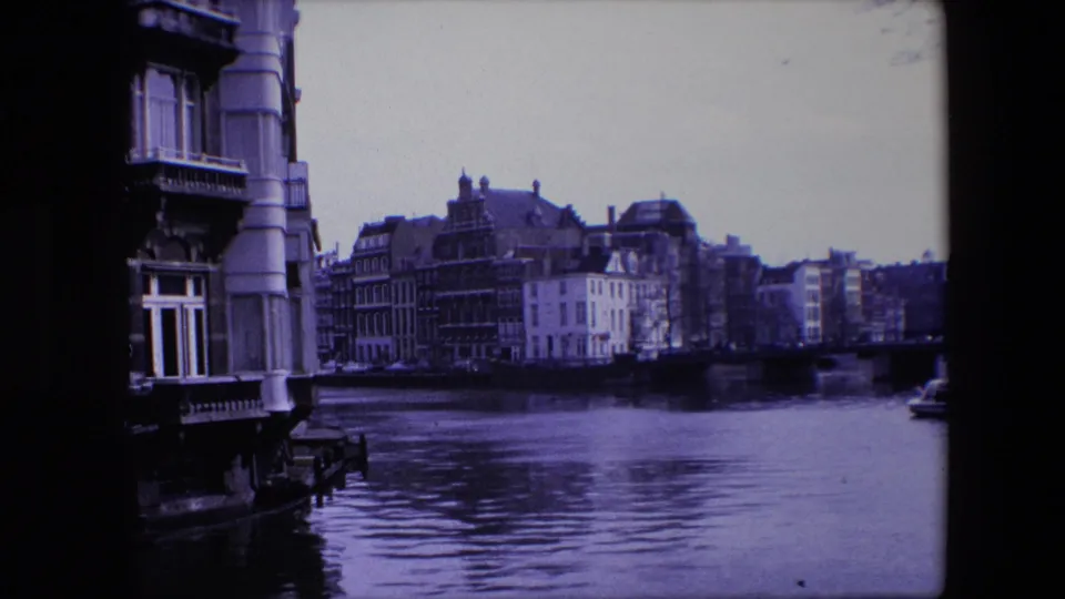 1967 Amsterdam canal scene with a close architectural edge, older waterfront buildings across the canal, and rippled water filling the foreground.