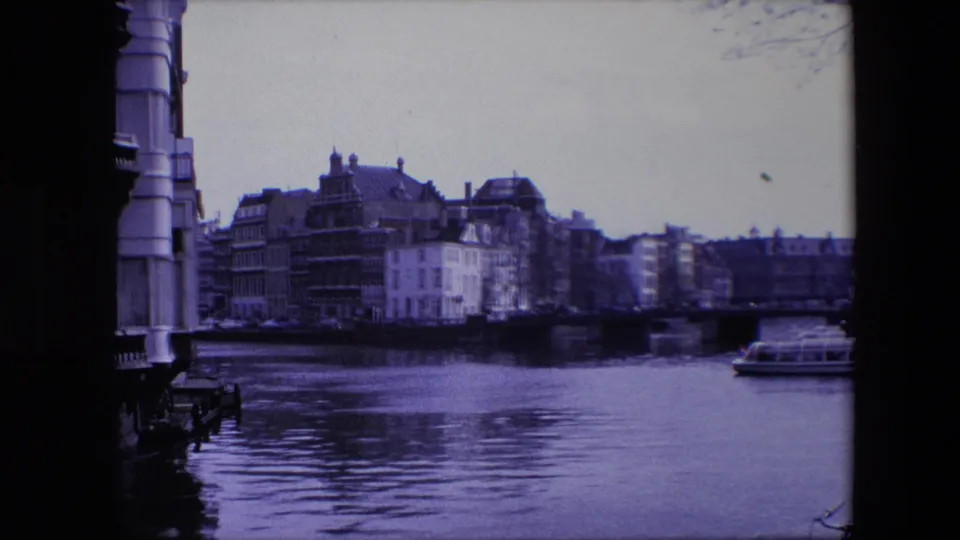 1967 Amsterdam canal view with historic buildings across the water and a small boat passing near the right side of the frame.