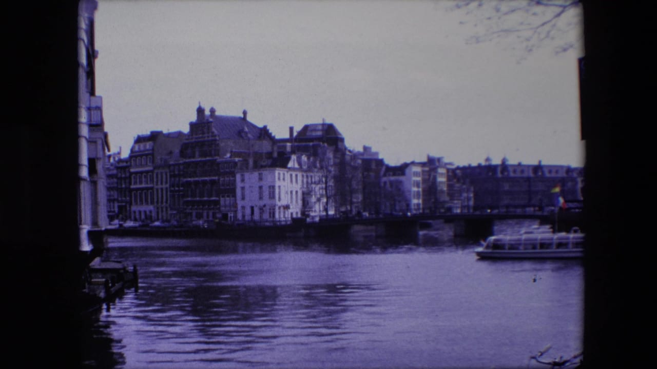 1967 8mm home-movie frame of an Amsterdam canal scene with waterfront buildings, a low bridge, rippled water, and a tour boat near the right side under a purple-blue film cast.