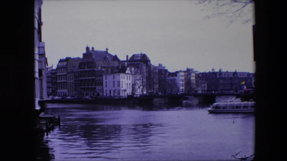 1967 8mm home-movie frame of an Amsterdam canal scene with waterfront buildings, a low bridge, rippled water, and a tour boat near the right side under a purple-blue film cast.