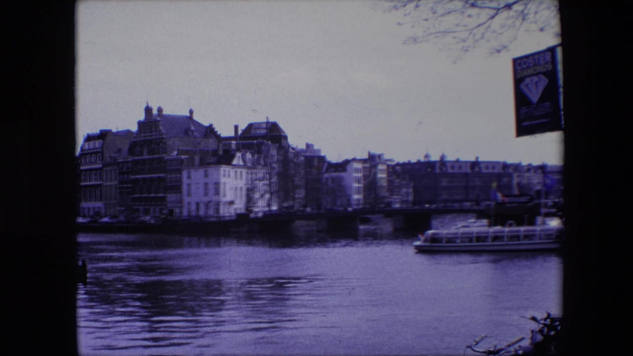 Amsterdam canal view from 1967 home-movie footage, showing historic-looking waterfront buildings, a bridge, tour boat, bare branches, and a Coster Diamonds sign near the upper right edge.