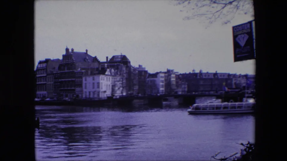 Amsterdam canal view from 1967 home-movie footage, showing historic-looking waterfront buildings, a bridge, tour boat, bare branches, and a Coster Diamonds sign near the upper right edge.