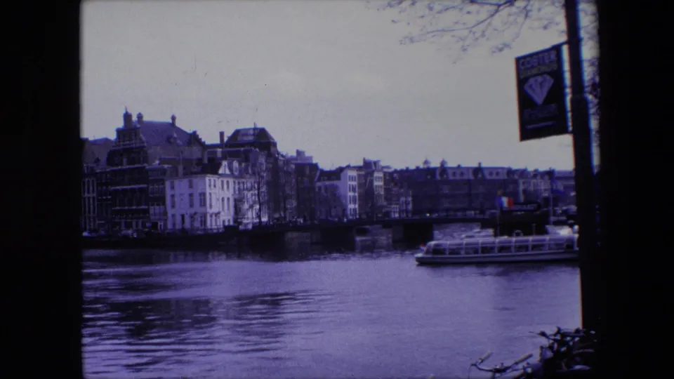 1967 Amsterdam 8mm frame with a purple cast, showing a canal, bridge, waterfront buildings, a tour boat passing along the right side, and a Coster Diamonds sign on a pole.