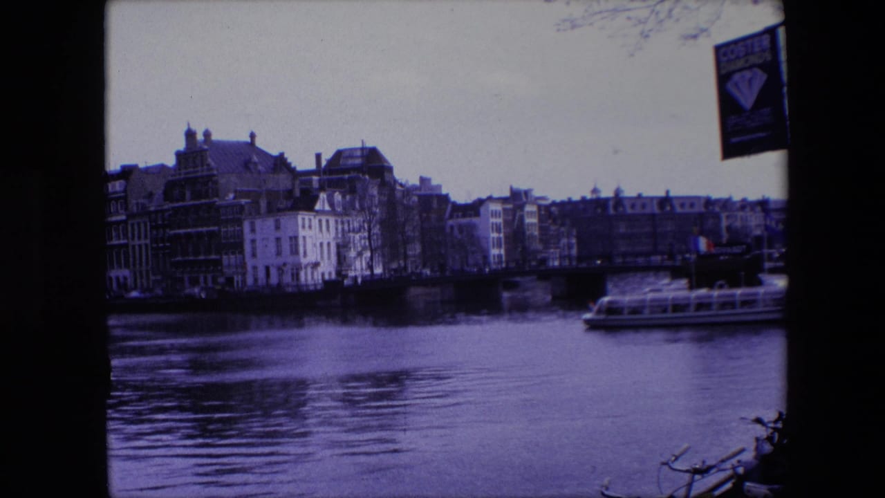 Final 1967 home-movie frame of the Amsterdam canal scene, with rippled water, a low bridge, dense waterfront buildings, a tour boat at the right, and visible film gate borders.