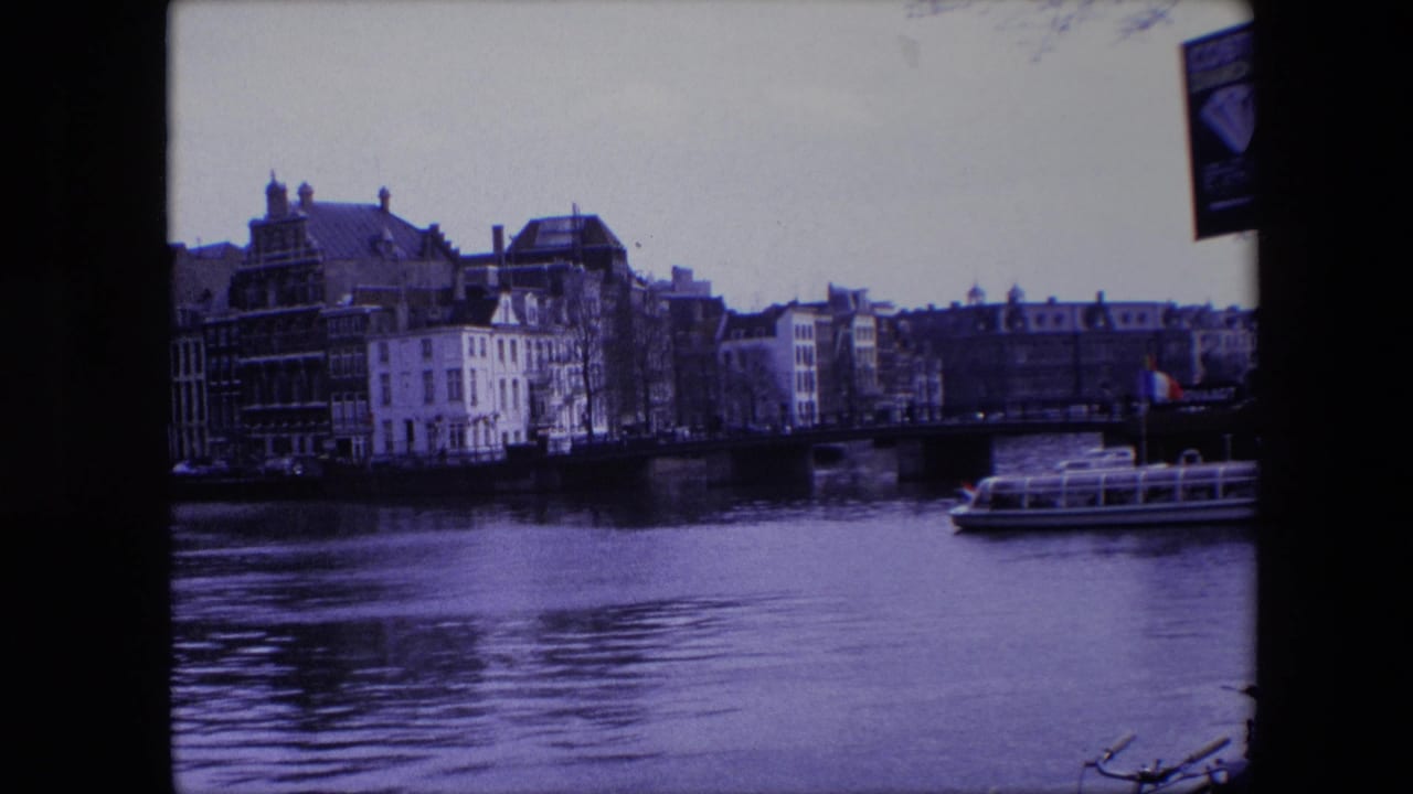 1967 Amsterdam canal view with multi-story buildings along the water, a low bridge, and a glass-roofed tour boat moving at the right edge.
