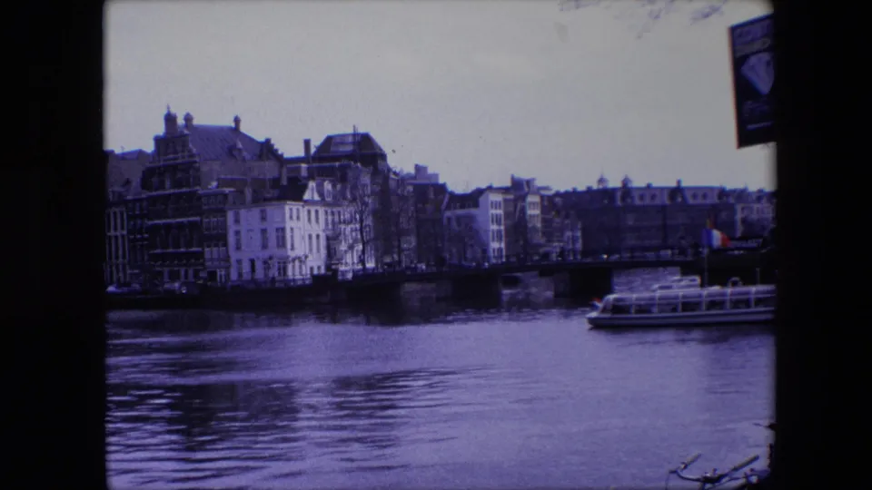 1967 Amsterdam canal view with multi-story buildings along the water, a low bridge, and a glass-roofed tour boat moving at the right edge.