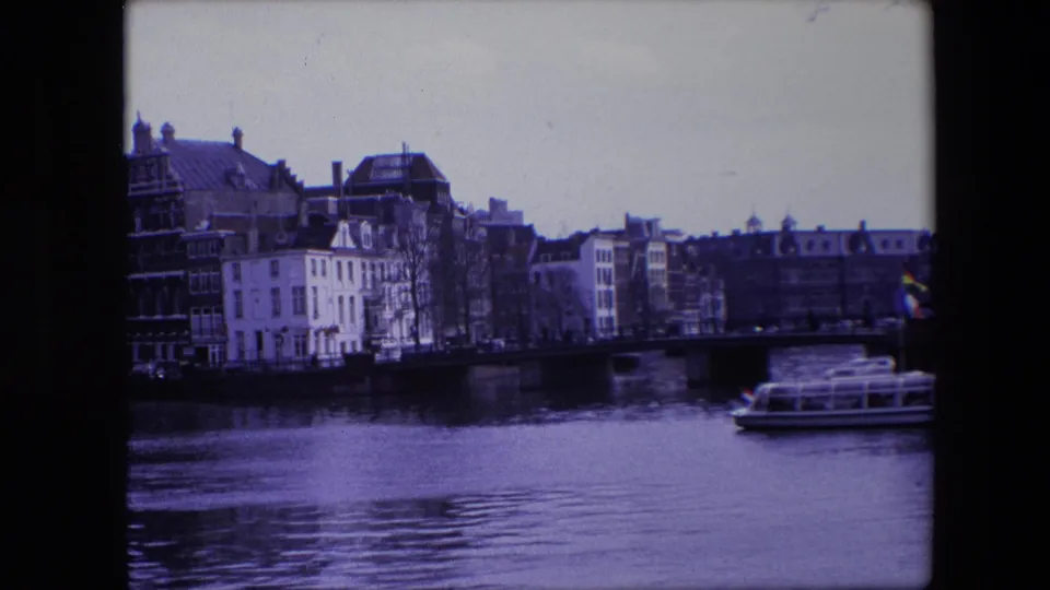 Amsterdam canal scene from the supplied 1967 footage, showing a tour boat beside historic-looking buildings and a bridge under a pale sky.