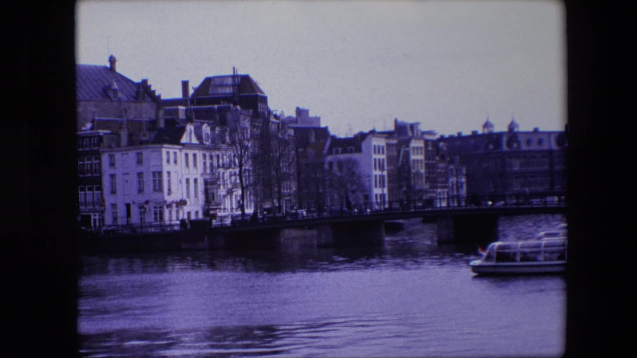 1967 Amsterdam canal view with a bridge, still water reflections, rows of buildings, and the front of a tour boat cropped at the lower right.