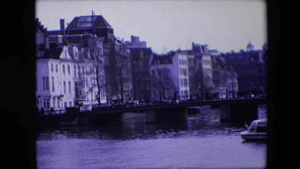Final frame from the 1967 Amsterdam canal clip, with waterfront architecture, a bridge over the canal, bare trees, and a cropped tour boat at right.