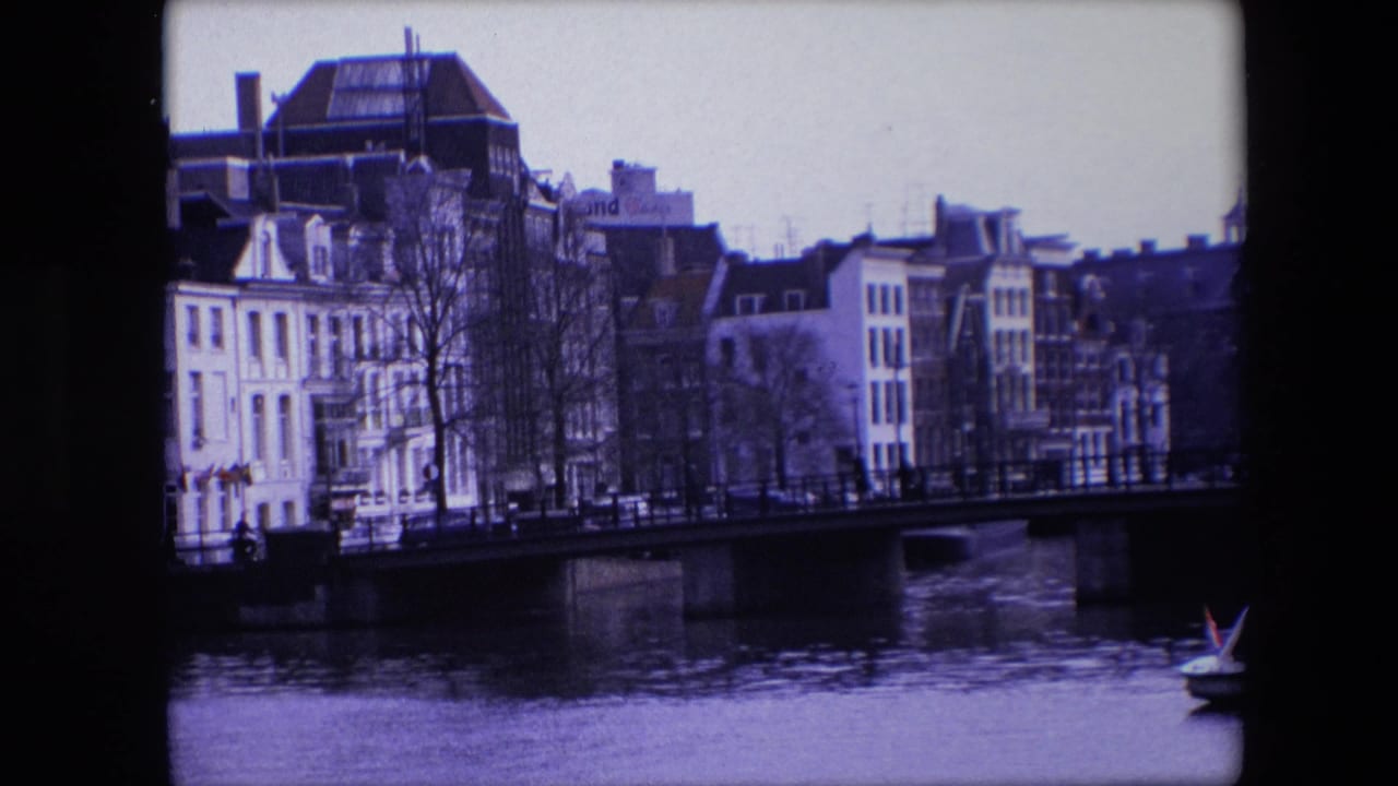 1967 Amsterdam canal view with a low bridge, narrow waterfront buildings, bare winter trees, and a small boat carrying a Dutch flag near the frame edge.