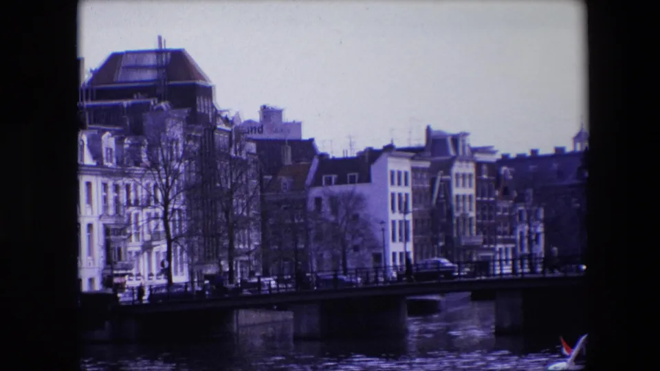 Amsterdam canal scene from 1967 showing a bridge in front of traditional buildings, bare trees along the water, small cars, and a boat with a Dutch flag at the edge.