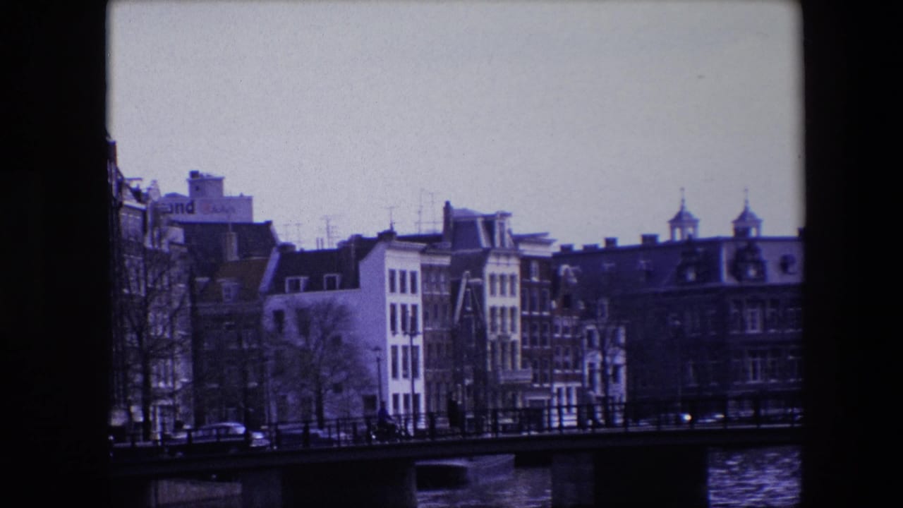 1967 Amsterdam canal-side buildings seen behind a low bridge, with bare trees, dark railings, cars on the street, and rippled water in the foreground.