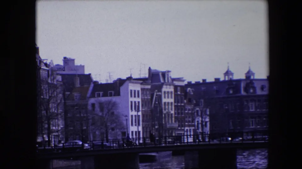 1967 Amsterdam canal-side buildings seen behind a low bridge, with bare trees, dark railings, cars on the street, and rippled water in the foreground.