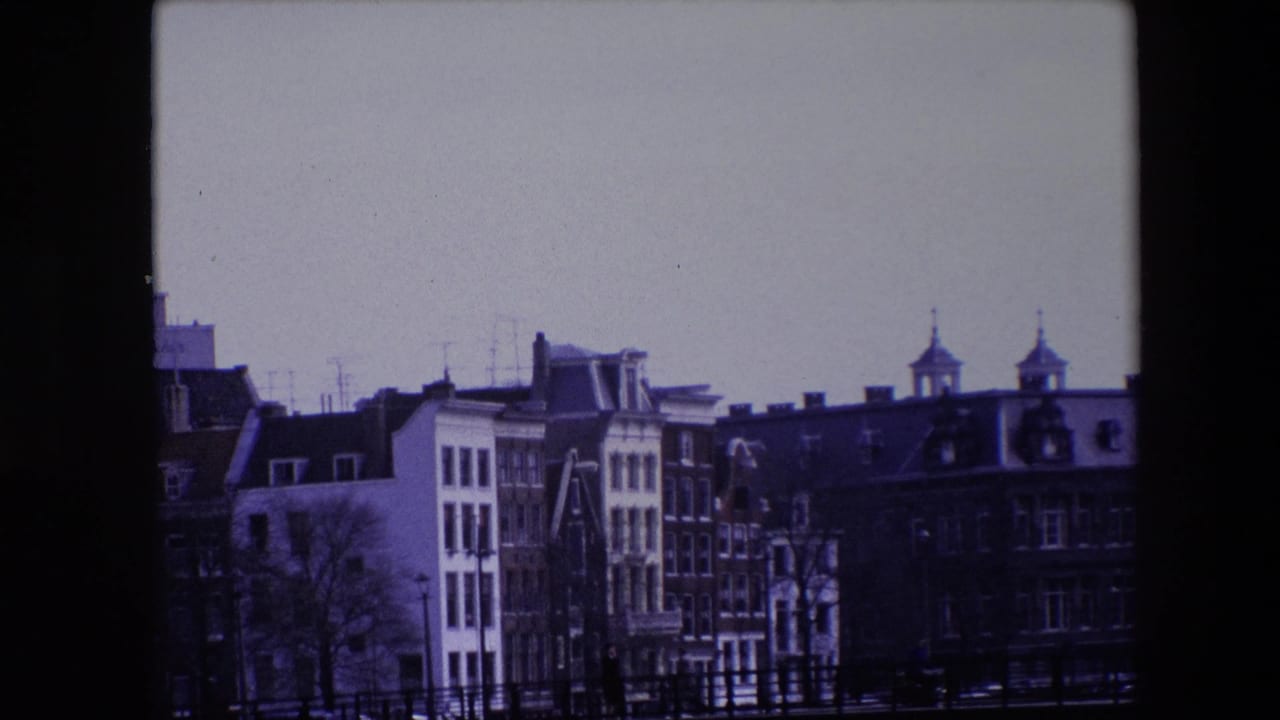 Amsterdam waterfront in 1967 with a bridge crossing the canal, rows of narrow buildings and rooflines beyond it, bare trees, railings, and dark water below.
