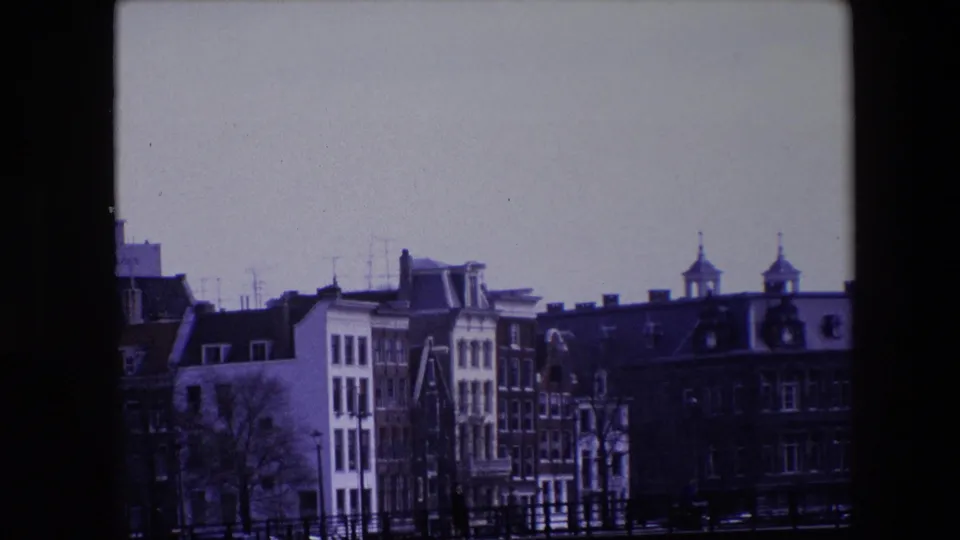 Amsterdam waterfront in 1967 with a bridge crossing the canal, rows of narrow buildings and rooflines beyond it, bare trees, railings, and dark water below.