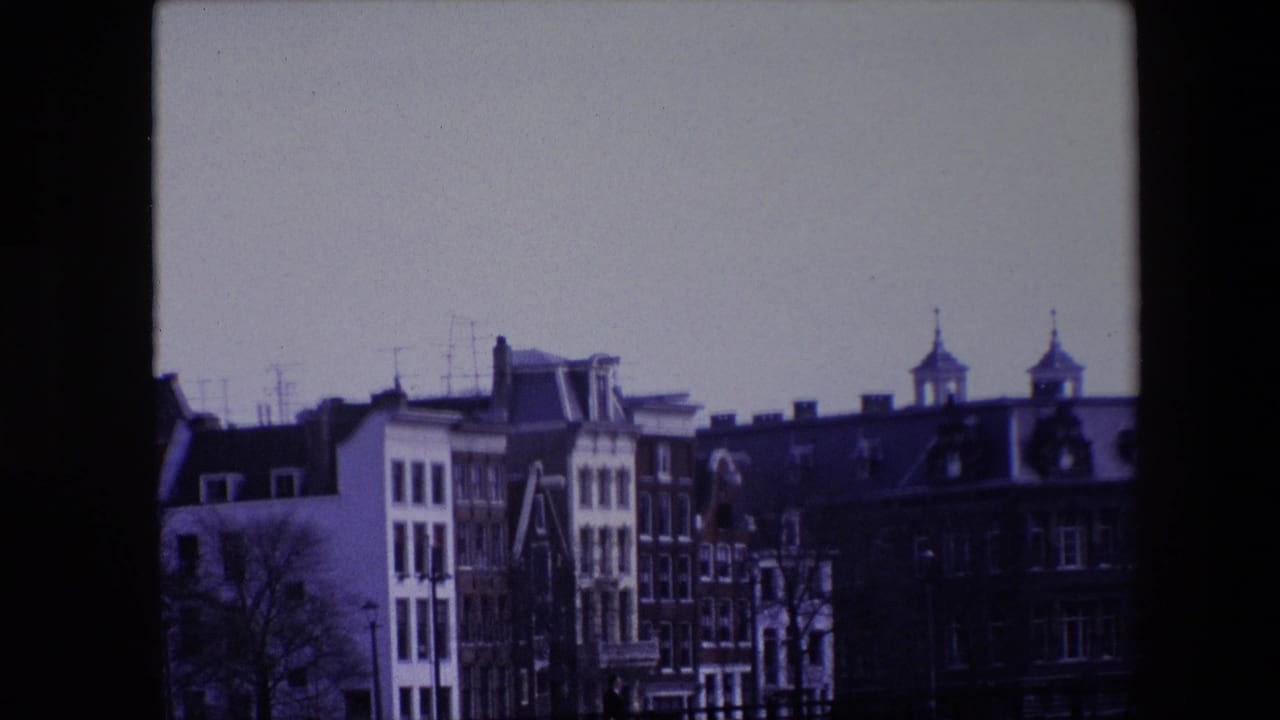 1967 8mm Amsterdam view showing traditional canal-house facades beneath a pale sky, with rooftop antennas and two spired towers in the background.