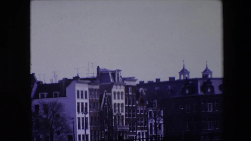 1967 8mm Amsterdam view showing traditional canal-house facades beneath a pale sky, with rooftop antennas and two spired towers in the background.