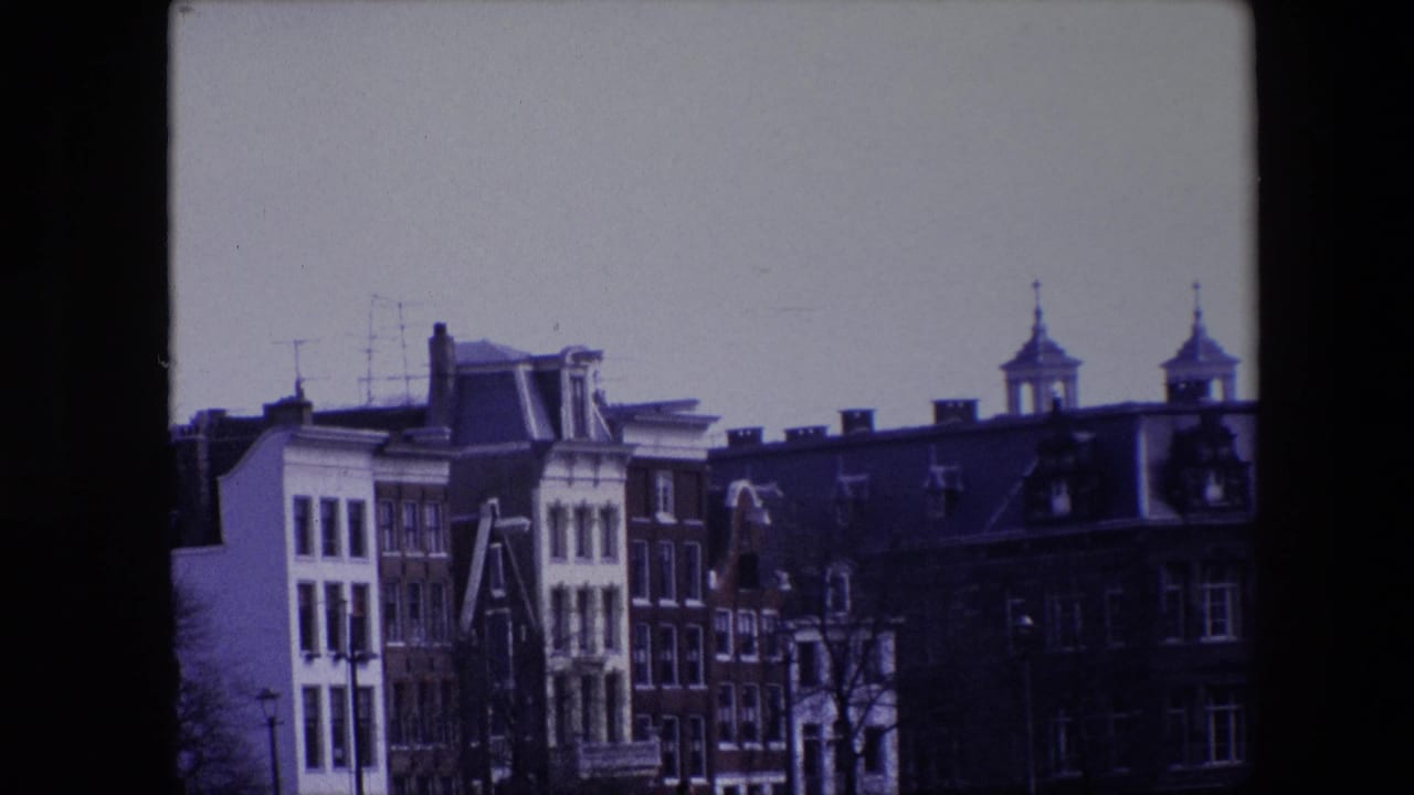 1967 Amsterdam home-movie frame with narrow historic buildings, dark rooflines, dormer windows, rooftop antennas, and two small towers above the skyline.