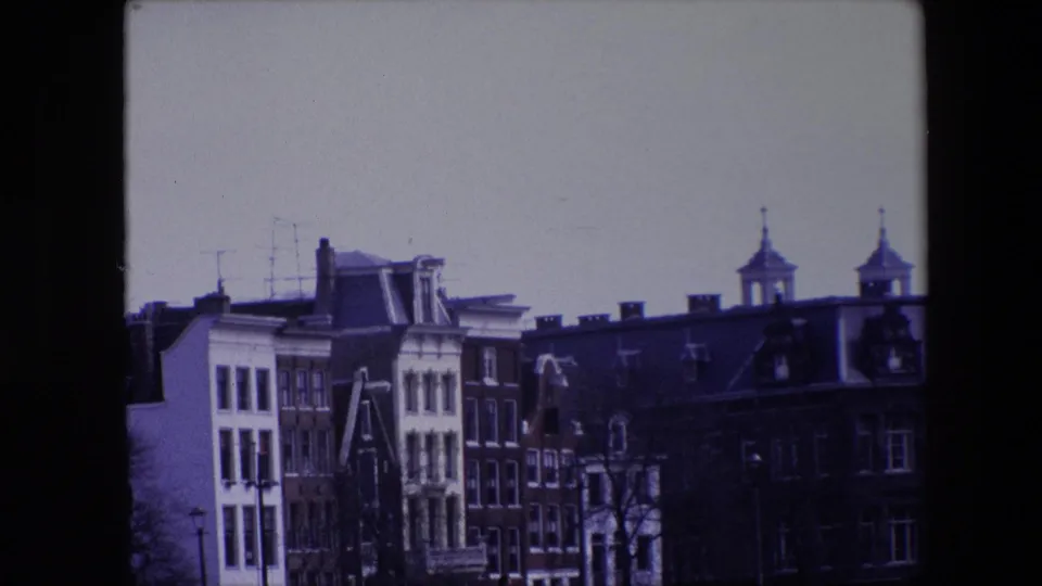 1967 Amsterdam home-movie frame with narrow historic buildings, dark rooflines, dormer windows, rooftop antennas, and two small towers above the skyline.