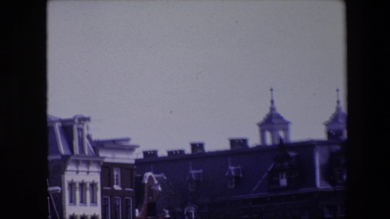 1967 8mm frame of Amsterdam rooftops during a pan, emphasizing dormers, chimneys, roof antennas, and two spired towers against a washed pale sky.