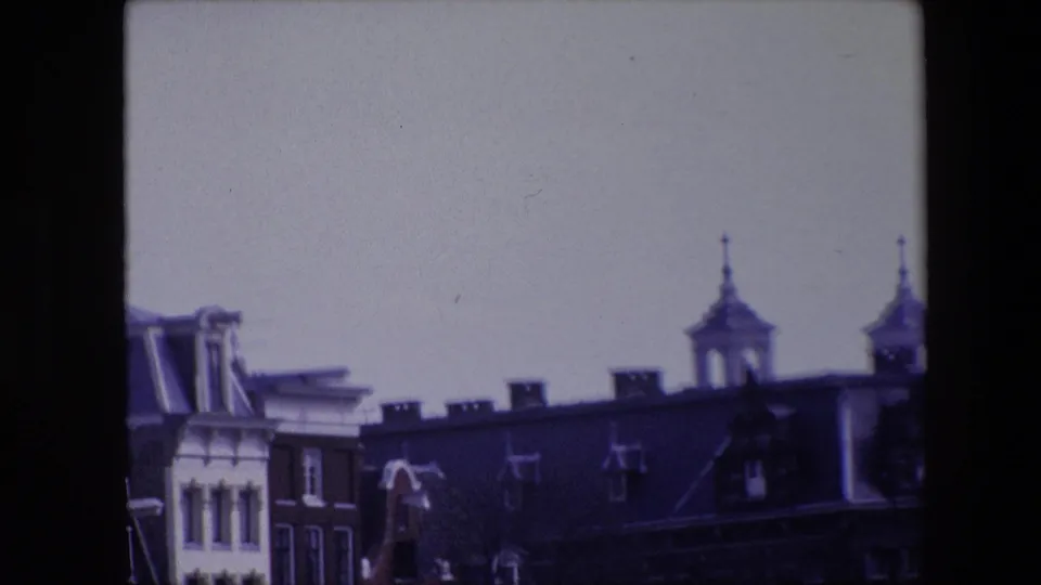 1967 8mm frame of Amsterdam rooftops during a pan, emphasizing dormers, chimneys, roof antennas, and two spired towers against a washed pale sky.