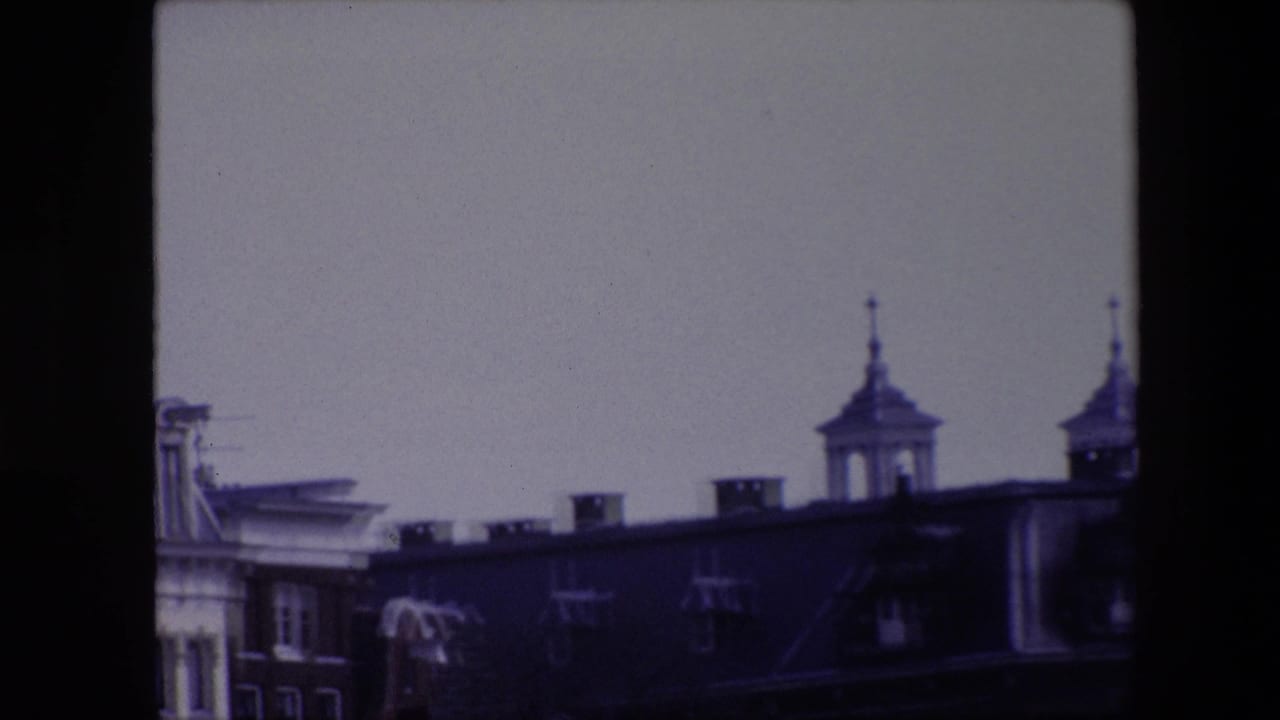 1967 Amsterdam home-movie frame showing a dark roofline, partial towered facade, pale sky, and pronounced 8mm frame edges.