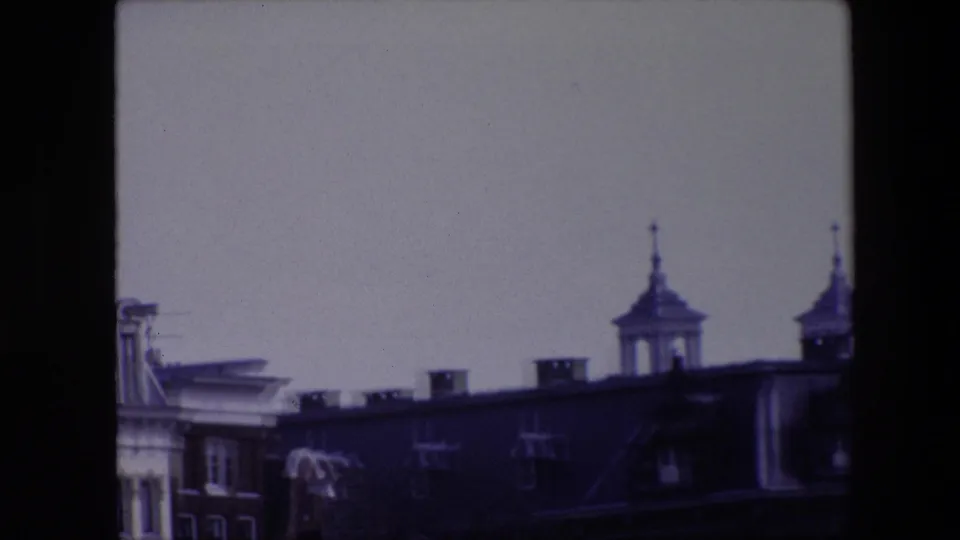 1967 Amsterdam home-movie frame showing a dark roofline, partial towered facade, pale sky, and pronounced 8mm frame edges.