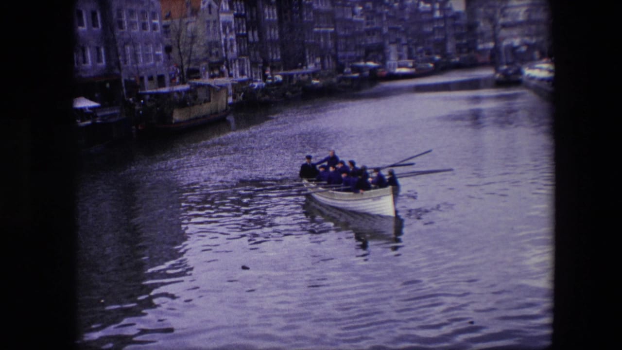 1967 Amsterdam canal view with a rowboat carrying several people, rippled water, waterfront buildings, moored boats, and parked cars along the canal edge.