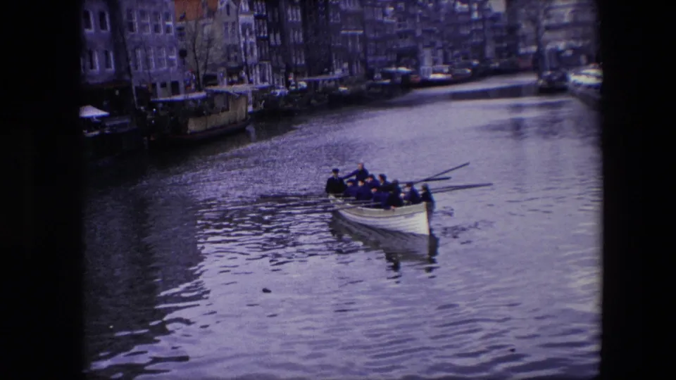 1967 Amsterdam canal view with a rowboat carrying several people, rippled water, waterfront buildings, moored boats, and parked cars along the canal edge.