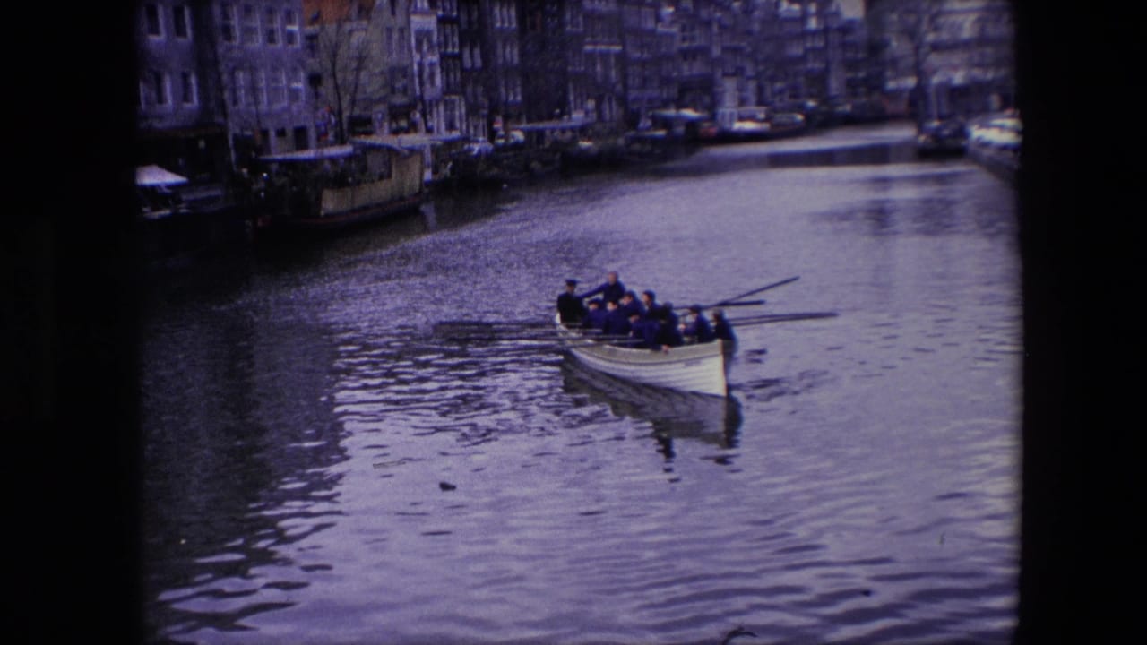 Adult men row a white boat along an Amsterdam canal in 1967, seen in soft 8mm footage with canal-front buildings in the background.