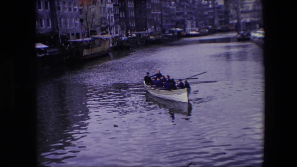 Adult men row a white boat along an Amsterdam canal in 1967, seen in soft 8mm footage with canal-front buildings in the background.