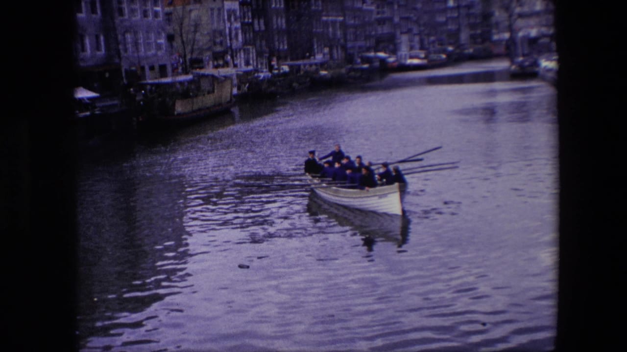 A rowing crew in dark clothing moves across an Amsterdam canal, with moored boats and narrow urban buildings visible behind them.