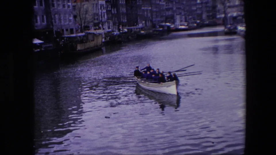 A rowing crew in dark clothing moves across an Amsterdam canal, with moored boats and narrow urban buildings visible behind them.