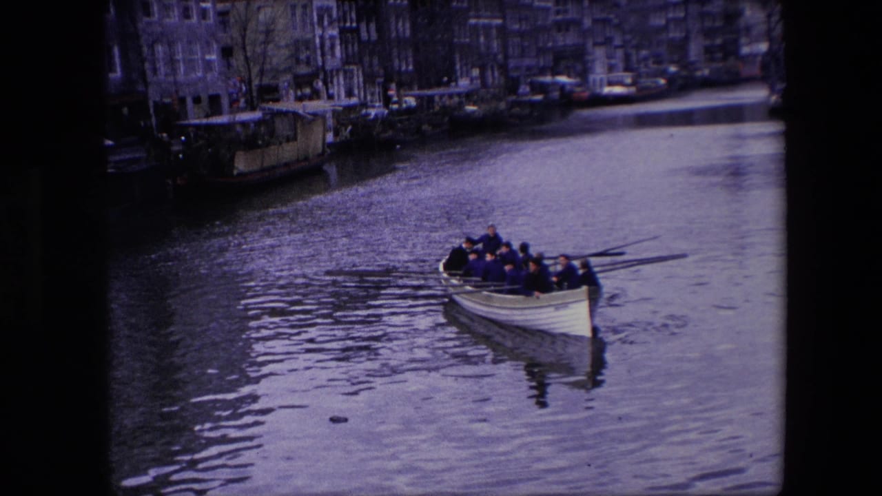 Several adult men in a white rowboat pass closer to the camera on an Amsterdam canal, surrounded by rippled water and soft 8mm grain.